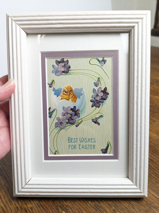 Postcard shows a hatching chick with flowers decorated with a light dusting of glitter. Text says "Best Wishes for Easter" on the bottom of the print. Framed in a vintage white frame to hang or set on a table top.