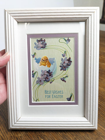Postcard shows a hatching chick with flowers decorated with a light dusting of glitter. Text says "Best Wishes for Easter" on the bottom of the print. Framed in a vintage white frame to hang or set on a table top.