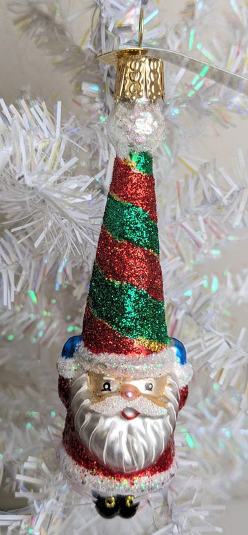 A smiling santa ornament made of blown glass and hand painted with his traditional red suit but a green and red striped hat. He's holding onto his hat with hands bundled up in blue mittens.