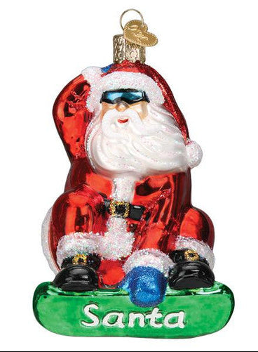 Sunglass wearing Santa holding onto his hat while he's shredding the snow. The name SANTA is emblazoned on his green snowboard.  His mittens are blue.